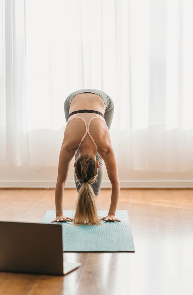 pexels-photo-6453407-6453407 Woman doing a standing forward bend yoga pose at home with a laptop on the floor.