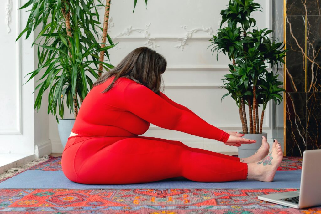 pexels-photo-6975648-6975648 Side view full body of plus size female in red activewear practicing Pascimottanasana posture during online yoga lesson on laptop at home